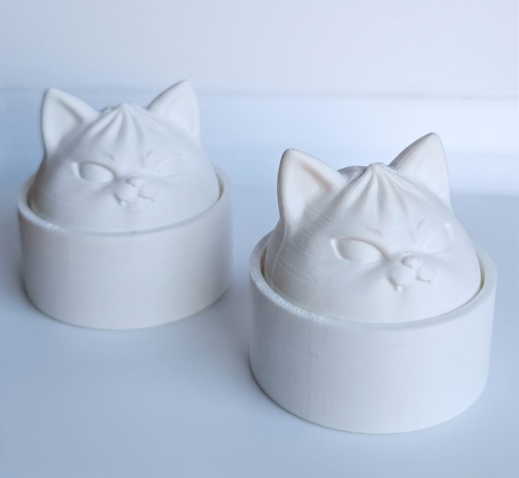 Kaks üleni valget 3D-prinditud kassikujulist detaili koos ümmarguste alustega heledal taustal, millel on näha prinditud materjali kihiline tekstuur ja varjud.