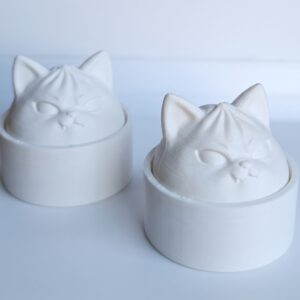 Kaks üleni valget 3D-prinditud kassikujulist detaili koos ümmarguste alustega heledal taustal, millel on näha prinditud materjali kihiline tekstuur ja varjud.